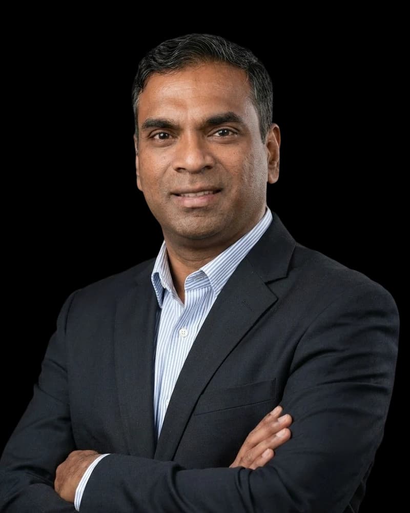 Dr. Pradeep Albert, smiling with his arms crossed, wearing a dark suit jacket over a blue and white striped shirt with an open collar.